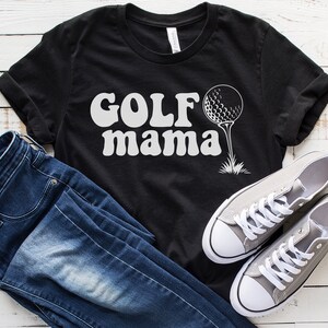 Op de afbeelding: Zwart T-shirt met witte tekst "GOLF mama" en een witte golfbal op een tee.