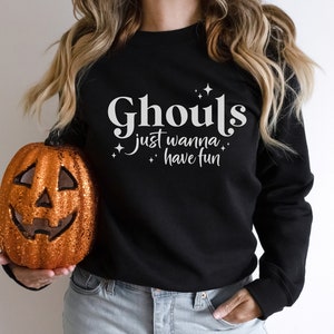 May include: Black sweatshirt with white text that reads "Ghouls just wanna have fun" with small white stars. A person is holding a glittery orange pumpkin.