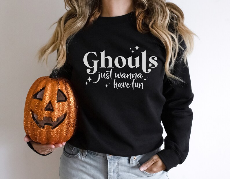 May include: Black sweatshirt with white text that reads "Ghouls just wanna have fun" with small white stars. A person is holding a glittery orange pumpkin.