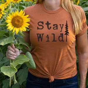 May include: A woman wearing a burnt orange t-shirt with the text "Stay Wild" and a small pine tree graphic. She is holding a bouquet of sunflowers in a field.