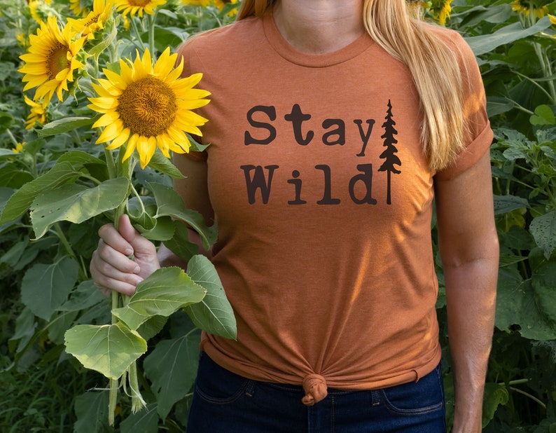May include: A woman wearing a burnt orange t-shirt with the text "Stay Wild" and a small pine tree graphic. She is holding a bouquet of sunflowers in a field.