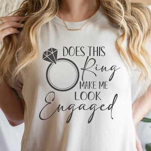 May include: White t-shirt with the text "Does this ring make me look engaged?" in black lettering. The text includes a stylized diamond ring graphic.