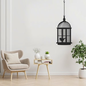 May include: A black silhouette of a bird in a hanging birdcage on a white wall. A beige armchair with a pillow sits in front of a small wooden table with white vases and plants.