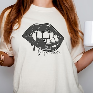 May include: A white t-shirt featuring a black graphic of a vampire mouth with fangs and the text "bite me".