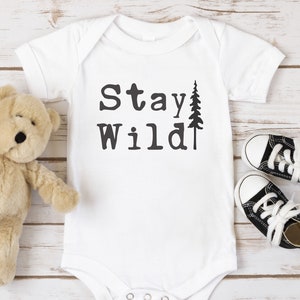 May include: White baby onesie with the text "Stay Wild" and a small pine tree graphic. A brown teddy bear and black and white baby shoes are on a white wood background.