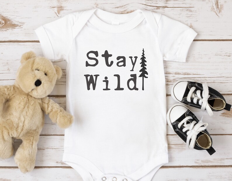 May include: White baby onesie with the text "Stay Wild" and a small pine tree graphic. A brown teddy bear and black and white baby shoes are on a white wood background.