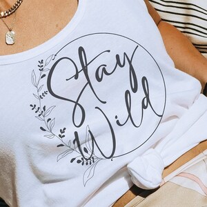 Puede incluir: Camiseta blanca de tirantes con un gráfico circular negro que dice "Stay Wild" con una corona de flores alrededor del círculo.