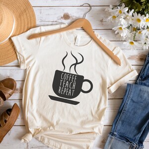 Może przedstawiać: Beżowa koszulka z czarnym nadrukiem przedstawiającym kubek kawy z parą unoszącą się nad nim. Tekst na kubku brzmi "Coffee Teach Repeat".