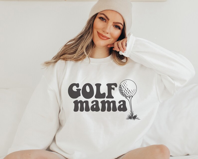 Op de afbeelding: Wit sweatshirt met de tekst "GOLF mama" in zwarte retro-stijl letters en een zwart-witte golfbal afbeelding.