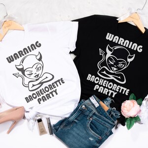 May include: Two white t-shirts with black text and graphics. The text on the shirts reads "Warning Bachelorette Party". The graphic is a cartoon of a devil with horns and a tail. One shirt is on a black hanger and the other is on a white hanger. The shirts are on a white surface with a pair of jeans, a pair of high heels, and a bottle of perfume.
