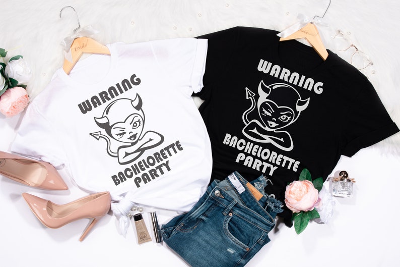 May include: Two white t-shirts with black text and graphics. The text on the shirts reads "Warning Bachelorette Party". The graphic is a cartoon of a devil with horns and a tail. One shirt is on a black hanger and the other is on a white hanger. The shirts are on a white surface with a pair of jeans, a pair of high heels, and a bottle of perfume.