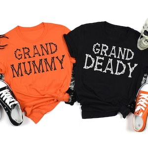 May include: Two black and orange t-shirts with a Halloween theme. The orange shirt says "Grand Mummy" and the black shirt says "Grand Deady".