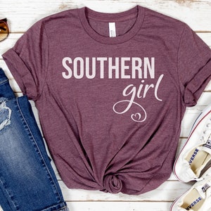 May include: A maroon t-shirt with the words "SOUTHERN girl" in white script. The shirt is styled with blue jeans, sunglasses, and white sneakers on a white wooden surface.