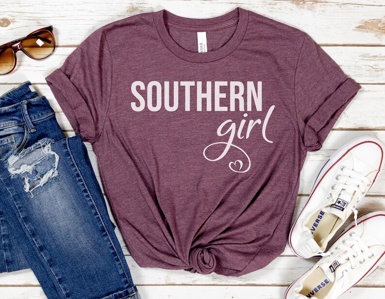 May include: A maroon t-shirt with the words "SOUTHERN girl" in white script. The shirt is styled with blue jeans, sunglasses, and white sneakers on a white wooden surface.