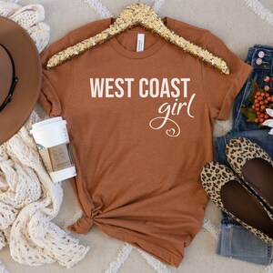 Puede incluir: Una camiseta de color &oacute;xido con el texto "WEST COAST girl" en letras blancas. La camiseta est&aacute; sobre una superficie con estampado blanco y marr&oacute;n con un sombrero marr&oacute;n, una taza de caf&eacute; blanca, unos vaqueros azules y un par de bailarinas de estampado de leopardo.