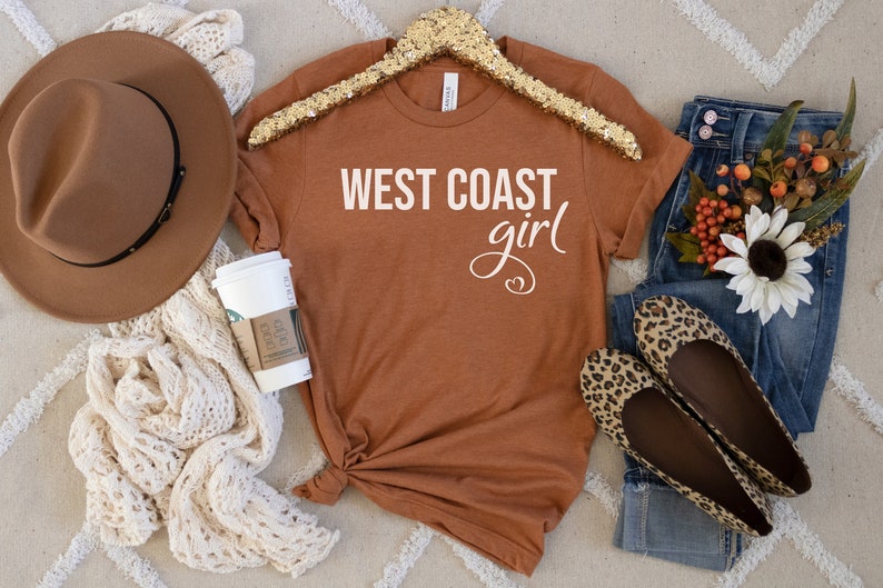 Puede incluir: Una camiseta de color &oacute;xido con el texto "WEST COAST girl" en letras blancas. La camiseta est&aacute; sobre una superficie con estampado blanco y marr&oacute;n con un sombrero marr&oacute;n, una taza de caf&eacute; blanca, unos vaqueros azules y un par de bailarinas de estampado de leopardo.