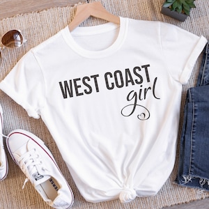 Puede incluir: Camiseta blanca con el texto "WEST COAST girl" en letras negras con un dise&ntilde;o de coraz&oacute;n.