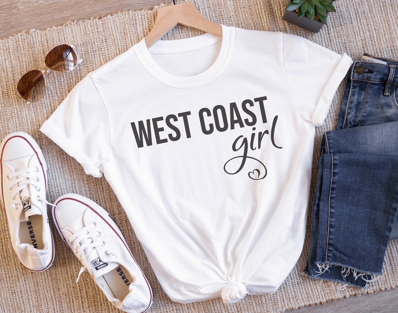 Puede incluir: Camiseta blanca con el texto "WEST COAST girl" en letras negras con un dise&ntilde;o de coraz&oacute;n.