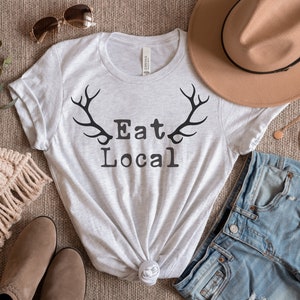 Puede incluir: Una camiseta gris con un gráfico negro de cuernos y el texto "Eat Local". La camiseta está colocada sobre una superficie marrón con un par de botas marrones, un sombrero marrón, unos pantalones cortos de mezclilla y un collar dorado.