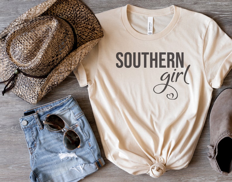 May include: A cream-colored t-shirt with the words "SOUTHERN girl" printed in black. The shirt is styled with denim shorts, sunglasses, a straw cowboy hat, and a pair of gray suede shoes. The shirt is knotted at the waist.