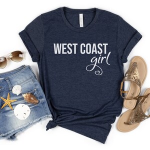 Puede incluir: Una camiseta azul marino con el texto "West Coast girl" en letras blancas. La camiseta se combina con pantalones cortos de mezclilla, sandalias y gafas de sol.