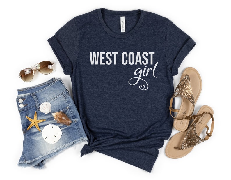 Puede incluir: Una camiseta azul marino con el texto "West Coast girl" en letras blancas. La camiseta se combina con pantalones cortos de mezclilla, sandalias y gafas de sol.