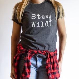 May include: A dark gray t-shirt with the text "Stay Wild" and a white silhouette of a pine tree. The shirt is worn by a person with a red and black plaid shirt tied around their waist.