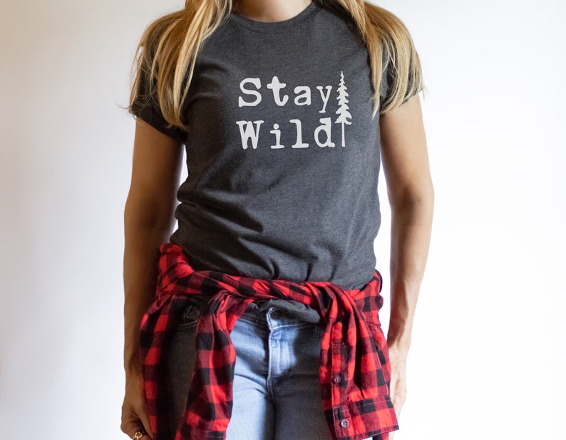 May include: A dark gray t-shirt with the text "Stay Wild" and a white silhouette of a pine tree. The shirt is worn by a person with a red and black plaid shirt tied around their waist.