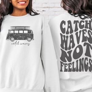 Puede incluir: Sudadera blanca con un gráfico de furgoneta vintage en la parte delantera y el texto "catch waves" debajo. La parte posterior de la sudadera tiene el texto "CATCH WAVES NOT FEELINGS" en una fuente retro.