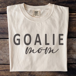 Pu&ograve; includere: T-shirt color crema con la scritta "GOALIE mom" stampata in grigio scuro. Il testo "GOALIE" &egrave; in grassetto e senza grazie, mentre "mom" &egrave; in corsivo. La maglietta ha un girocollo ed &egrave; realizzata in materiale morbido.