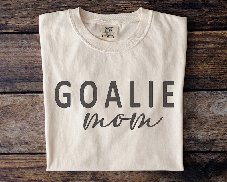 Pu&ograve; includere: T-shirt color crema con la scritta "GOALIE mom" stampata in grigio scuro. Il testo "GOALIE" &egrave; in grassetto e senza grazie, mentre "mom" &egrave; in corsivo. La maglietta ha un girocollo ed &egrave; realizzata in materiale morbido.