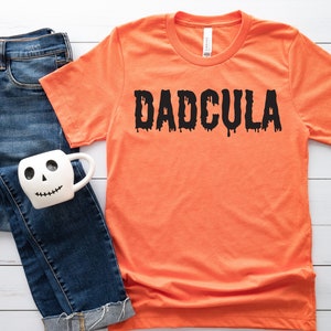 May include: Orange t-shirt with black dripping text that reads "DADCULA". The shirt is laying on a white wooden surface with a pair of blue jeans and a white mug with a skull face.