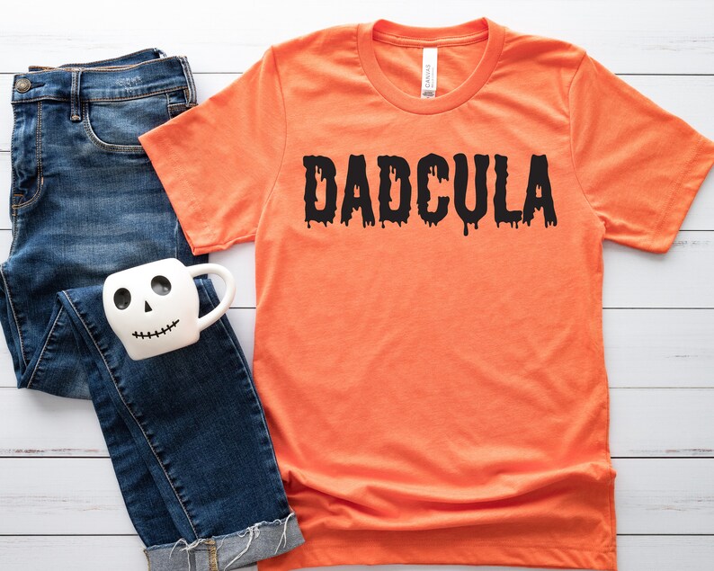 May include: Orange t-shirt with black dripping text that reads "DADCULA". The shirt is laying on a white wooden surface with a pair of blue jeans and a white mug with a skull face.