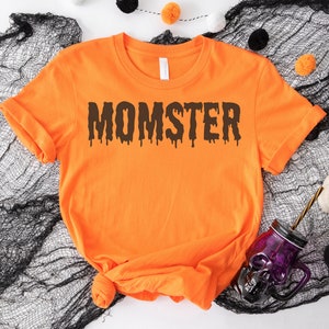 May include: Orange t-shirt with the text "MOMSTER" in black dripping letters. The shirt is on a black mesh background.