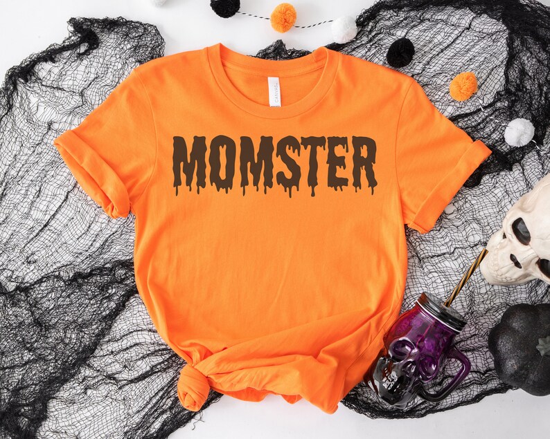 May include: Orange t-shirt with the text "MOMSTER" in black dripping letters. The shirt is on a black mesh background.