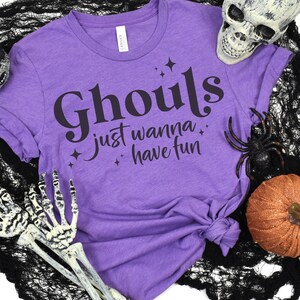 May include: Purple t-shirt with the text "Ghouls just wanna have fun" in black lettering with stars. The shirt is on a black and white background with a skull, spider, and pumpkin.