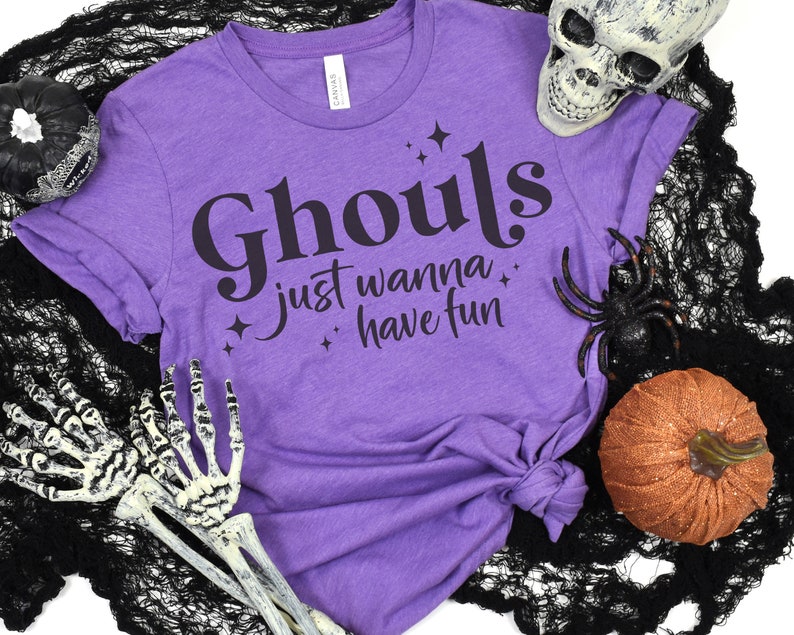 May include: Purple t-shirt with the text "Ghouls just wanna have fun" in black lettering with stars. The shirt is on a black and white background with a skull, spider, and pumpkin.