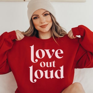 May include: A red sweatshirt with the words "love out loud" in white script. The sweatshirt is a crew neck style. The person in the image is wearing a beige beanie.