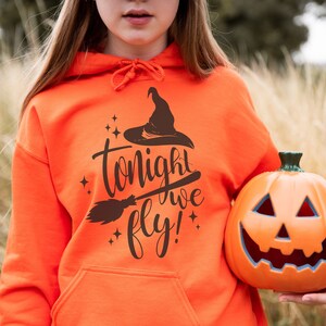 May include: A bright orange hoodie with a black witch hat and broom graphic and the text "tonight we fly!" A person is holding an orange jack-o'-lantern.