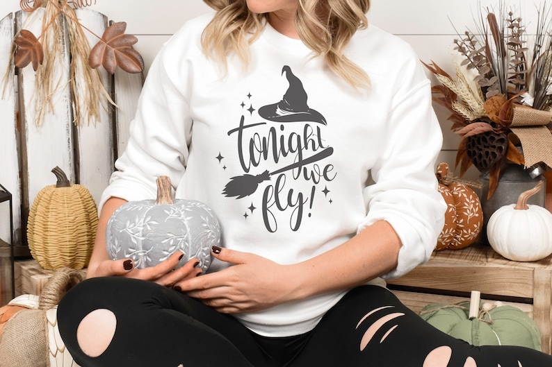 May include: A white sweatshirt with a black graphic design that says "Tonight we fly!" with a witch's hat and broom. The model is wearing black leggings and holding a grey pumpkin.