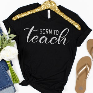 以下が含まれることがあります： シルバーの文字で「Born to teach」と書かれた黒いTシャツです。