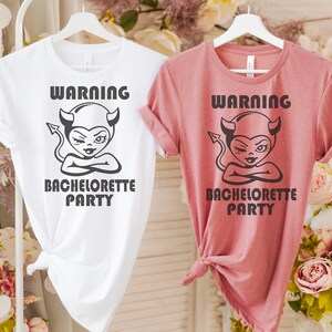 May include: Two t-shirts with a warning graphic of a devil with horns and a tail. The text on the shirts reads "Warning Bachelorette Party". One shirt is white and the other is pink.