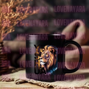 K&ouml;nnte beinhalten: Schwarze Keramik-Tasse mit einer L&ouml;wenkopf-Illustration in Gold- und Oranget&ouml;nen und dem Wort "YESHUA" in Gold. Die Tasse hat einen schwarzen Henkel und eine gl&auml;nzende Oberfl&auml;che. Ideal f&uuml;r Kaffee oder Tee.