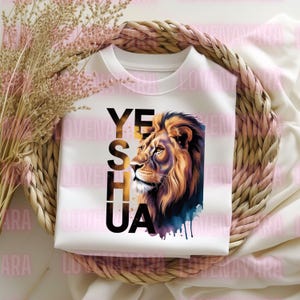 K&ouml;nnte beinhalten: Wei&szlig;es Sweatshirt mit einer L&ouml;wenillustration und dem Wort "YESHUA" in Schwarz und Gold. Die L&ouml;wenm&auml;hne ist detailliert mit Braun- und Goldt&ouml;nen und einem blauen Aquarelleffekt. Das Sweatshirt ist in einem geflochtenen Korb gefaltet.