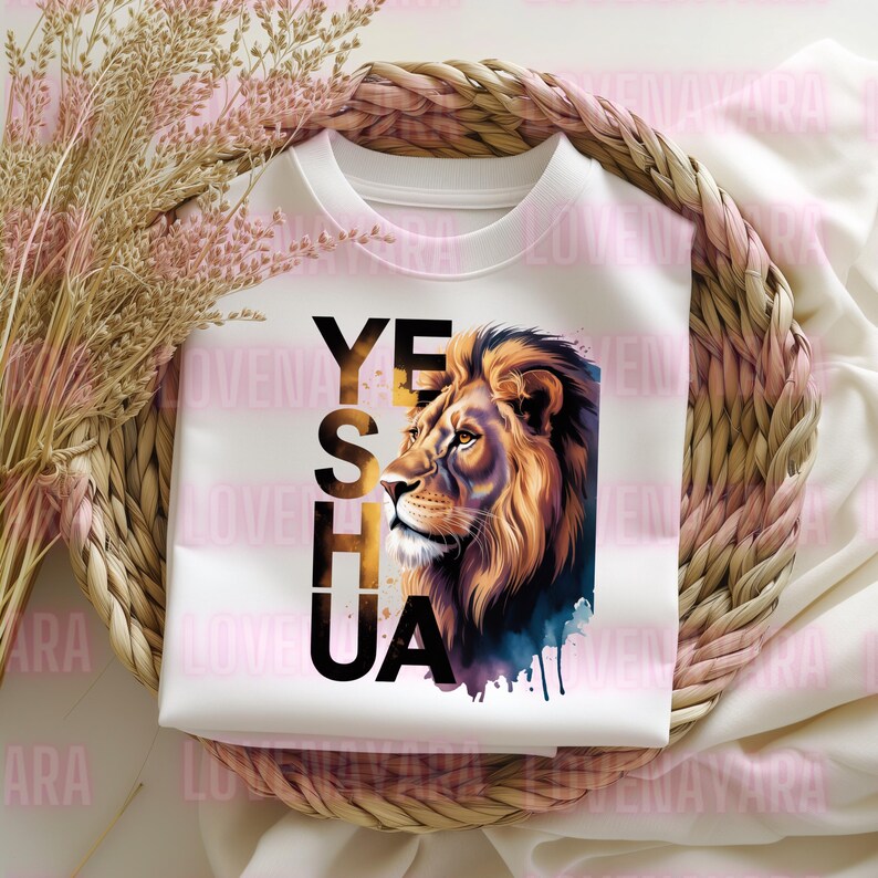 K&ouml;nnte beinhalten: Wei&szlig;es Sweatshirt mit einer L&ouml;wenillustration und dem Wort "YESHUA" in Schwarz und Gold. Die L&ouml;wenm&auml;hne ist detailliert mit Braun- und Goldt&ouml;nen und einem blauen Aquarelleffekt. Das Sweatshirt ist in einem geflochtenen Korb gefaltet.