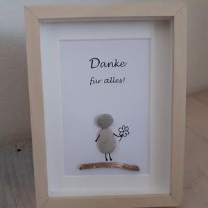 Könnte beinhalten: Ein gerahmtes Kunstwerk mit einer Person aus Kieselsteinen, die eine Blume hält, mit dem Text "Danke fur alles!" auf weißem Hintergrund.