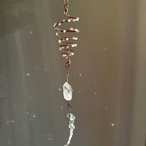 Op de afbeelding: Een hangend ornament met een koperkleurige draadspiraal versierd met kleine witte kralen. Onder de spiraal hangt een helder, ruw geslepen kristal aan een metalen schakel, gevolgd door een gebogen streng van iriserende, gefacetteerde glaskralen.