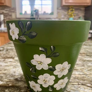 May include: A green terracotta flower pot with white painted flowers and green leaves. The pot is sitting on a light brown surface.