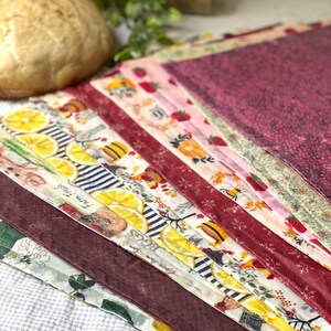 May include: A stack of rectangular fabric cloths featuring various patterns including yellow lemon slices, red strawberries, bees, and text reading Farm Fresh and Eggs. The cloths are layered in a diagonal arrangement with colors of maroon, white, and green.