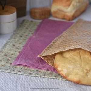 May include: A loaf of bread partially wrapped in a tan cloth, with a pink cloth and a floral patterned cloth underneath. The bread is golden brown and the cloths are folded neatly. The background includes jars and a wooden container.